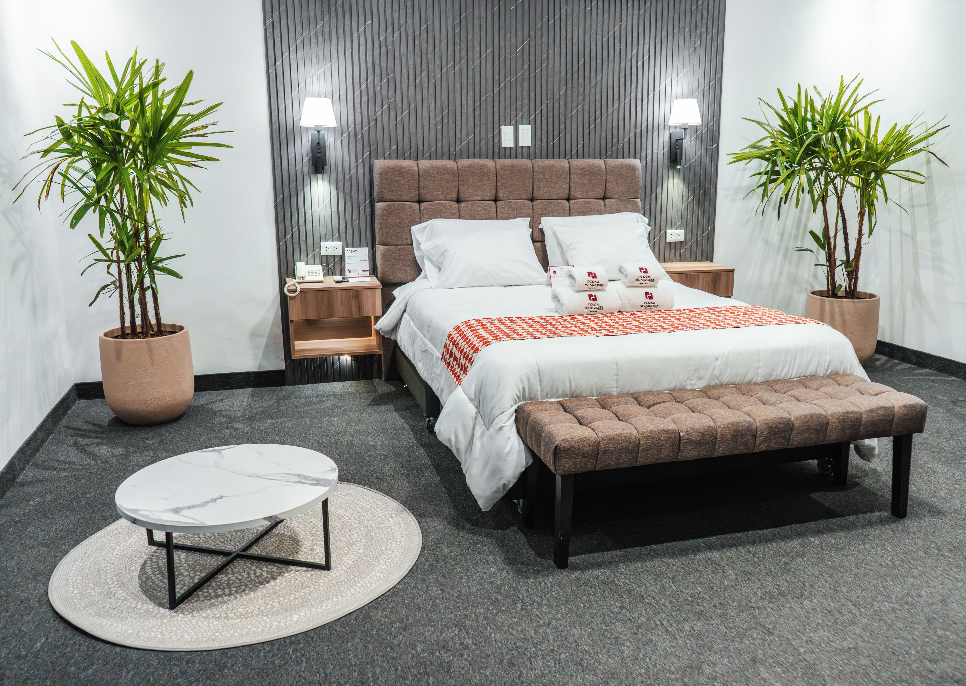 Una habitación de hotel moderna con una cama grande, mesitas de noche, una mesa de centro redonda y dos plantas en macetas.