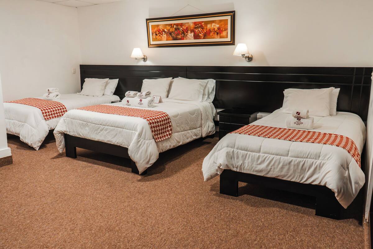 A hotel room with three single beds, each with a white comforter and red and white checkered blanket. There are two wall-mounted lamps on either side of the beds, and a framed abstract painting above the headboard.