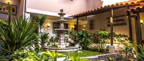 A courtyard with a central fountain, surrounded by lush greenery and a building with a tiled roof.