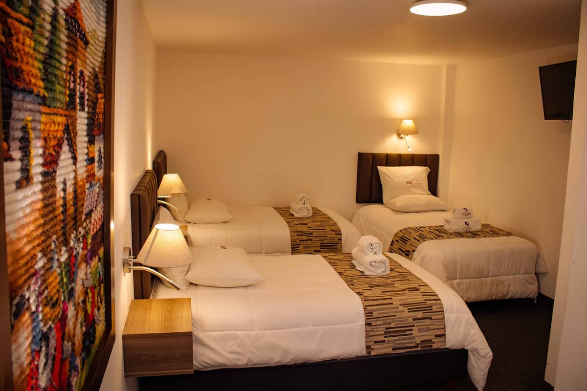 A hotel room with three beds, each with a headboard, a bedside lamp, and a small wooden nightstand.