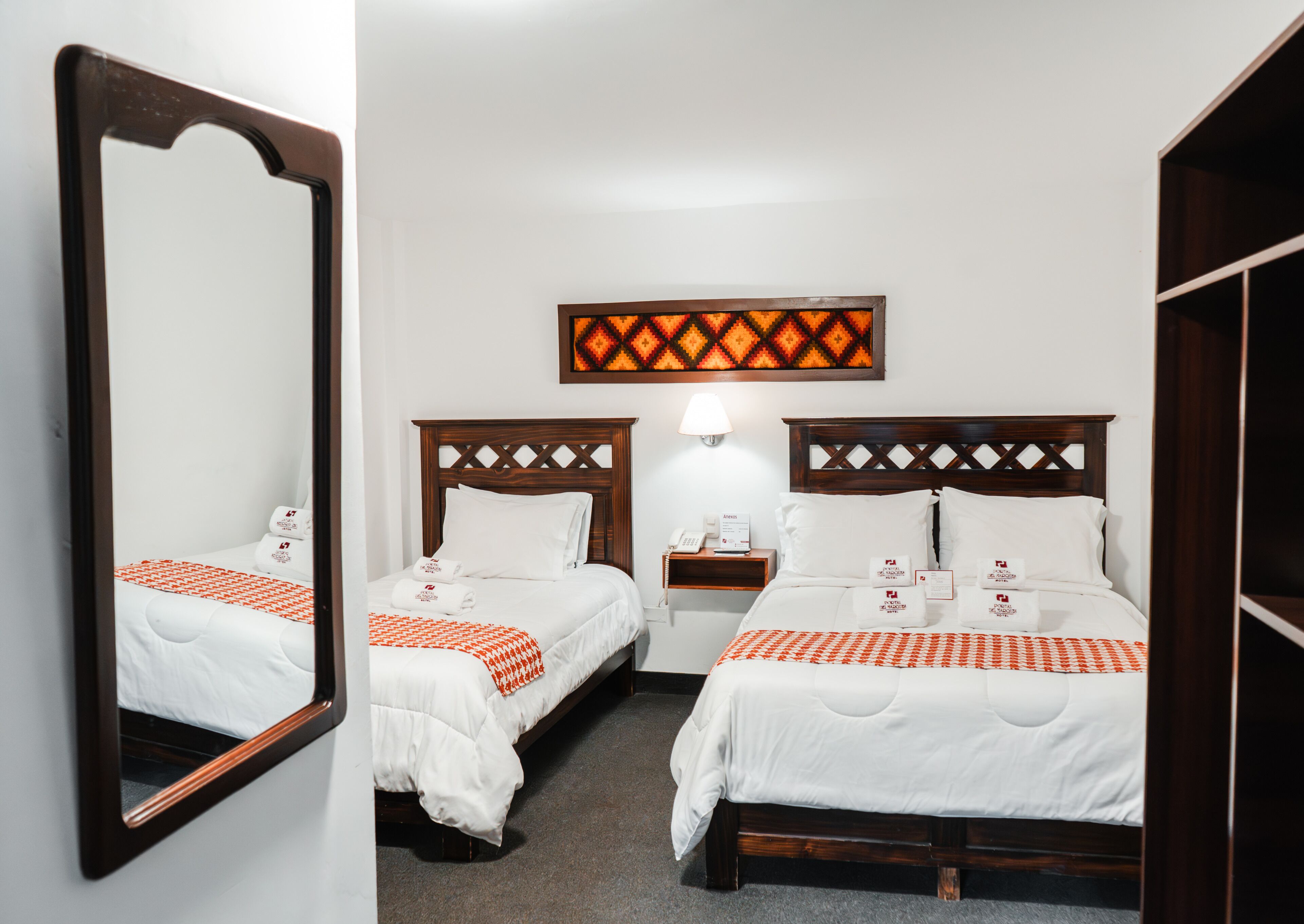 A hotel room with two beds, a nightstand with a lamp, a mirror, and a wall-mounted artwork.