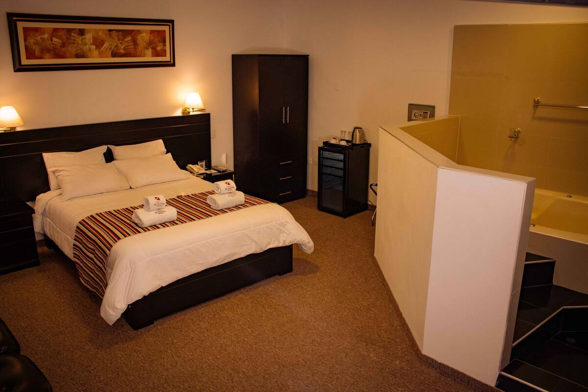 A hotel room with a bed, a wardrobe, a desk, a nightstand, a bathroom with a bathtub, and a framed picture on the wall.