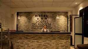 Una recepción con fondo de pared de piedra, un cartel que dice "HOTEL PORTAL DEL MARQUES CAIMARCO" y una recepcionista.