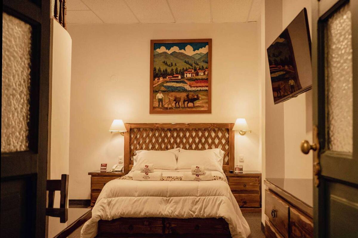 A hotel room with a wooden headboard, a bed with white bedding, two bedside lamps, and a painting on the wall.