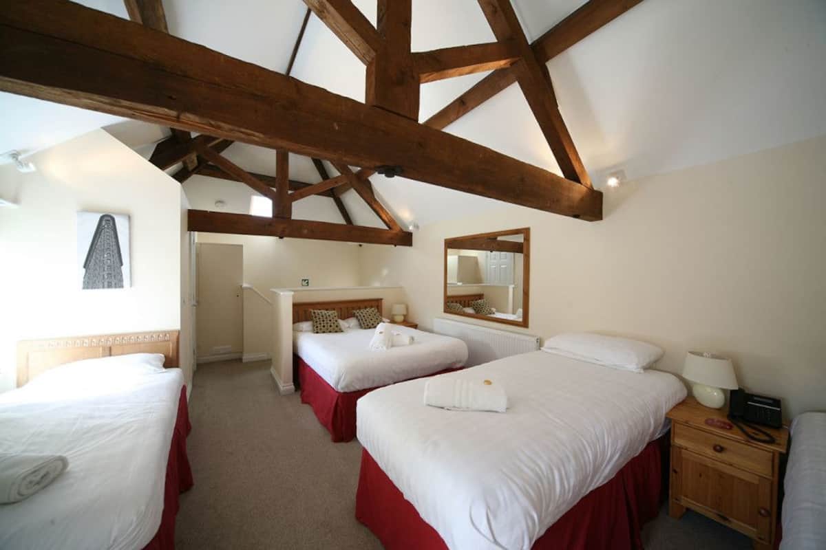 A room with three beds, wooden beams on the ceiling, a mirror, and a bedside table with a lamp.