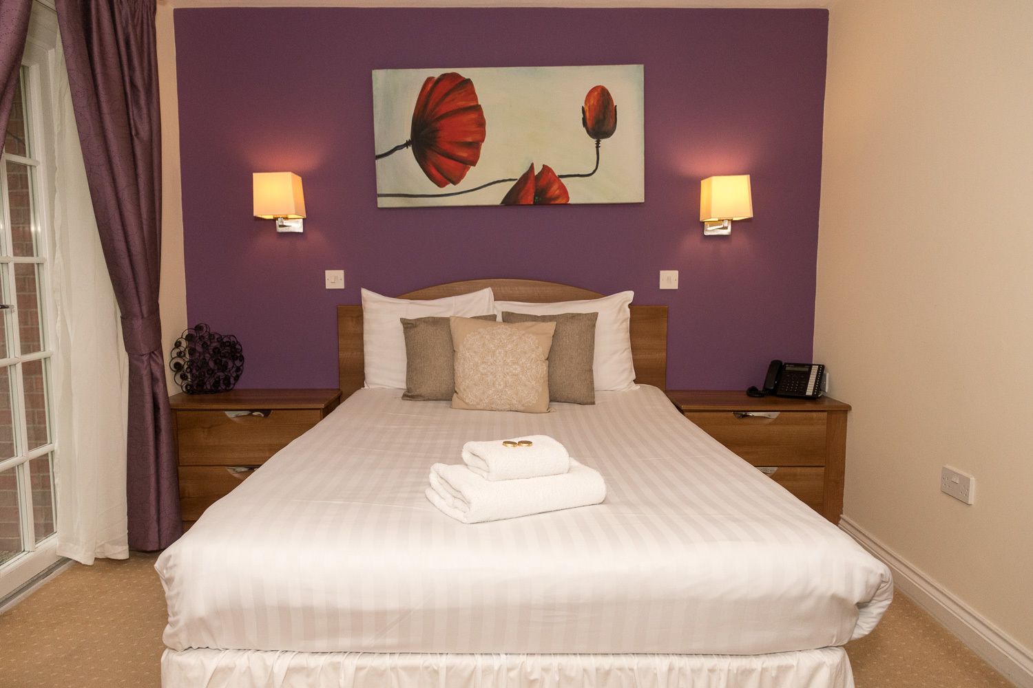 Una cama bien hecha con sábanas blancas y una toalla doblada, un cabecero de madera, dos mesitas de noche con lámparas, y un cuadro de flores rojas en la pared.