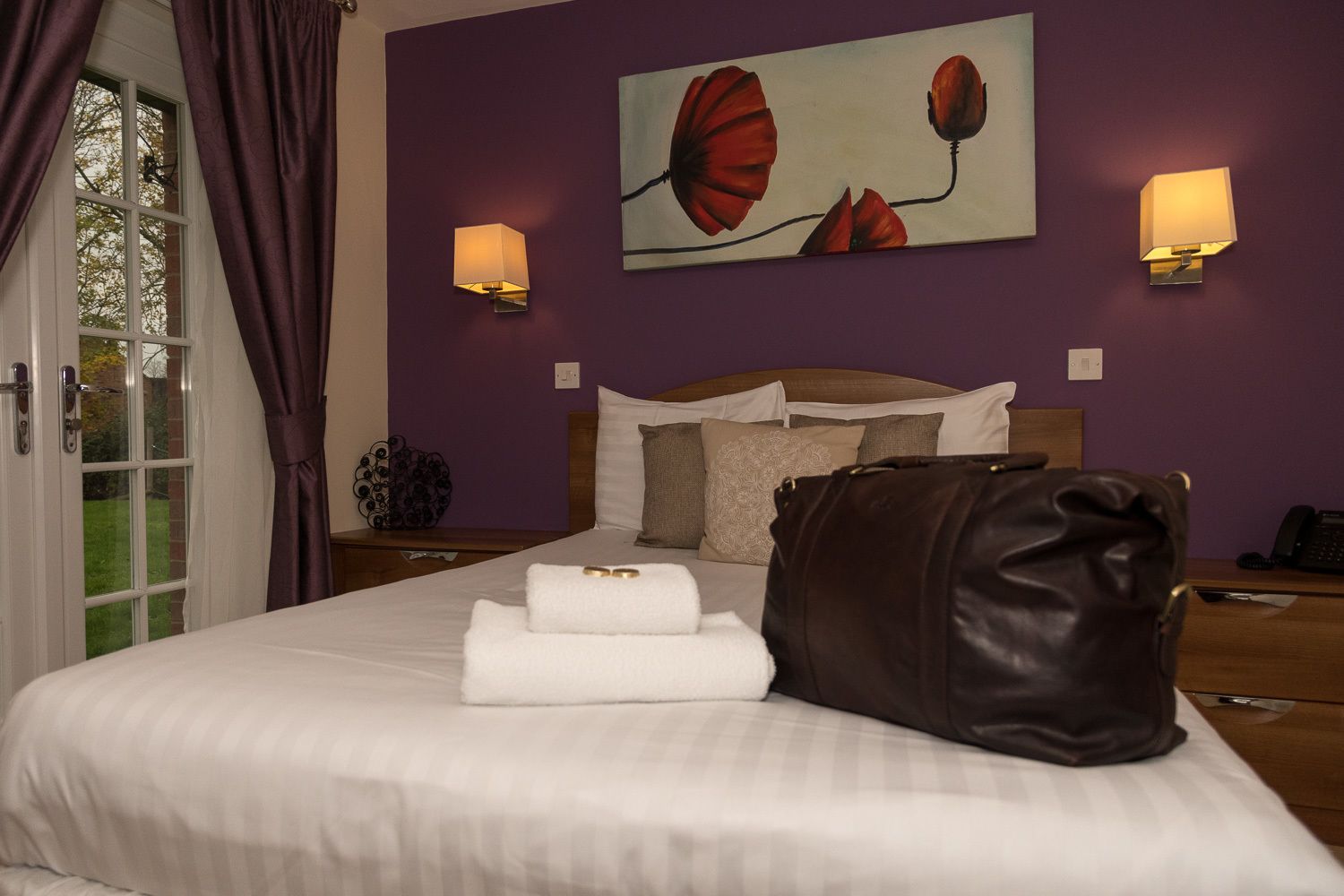A neatly made bed with white linens, a brown leather pillow, and a folded towel. There are two wall-mounted lamps on either side of a painting of red flowers.