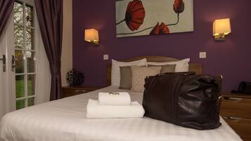 A neatly made bed with white linens, a brown leather pillow, and a folded towel. There are two wall-mounted lamps on either side of a painting of red flowers.