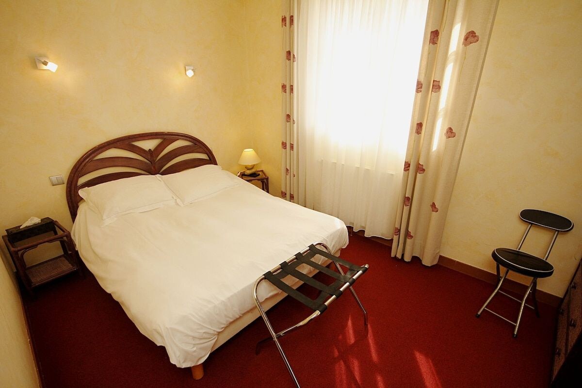 A hotel room with a bed, a nightstand, a lamp, a window with curtains, and two chairs.