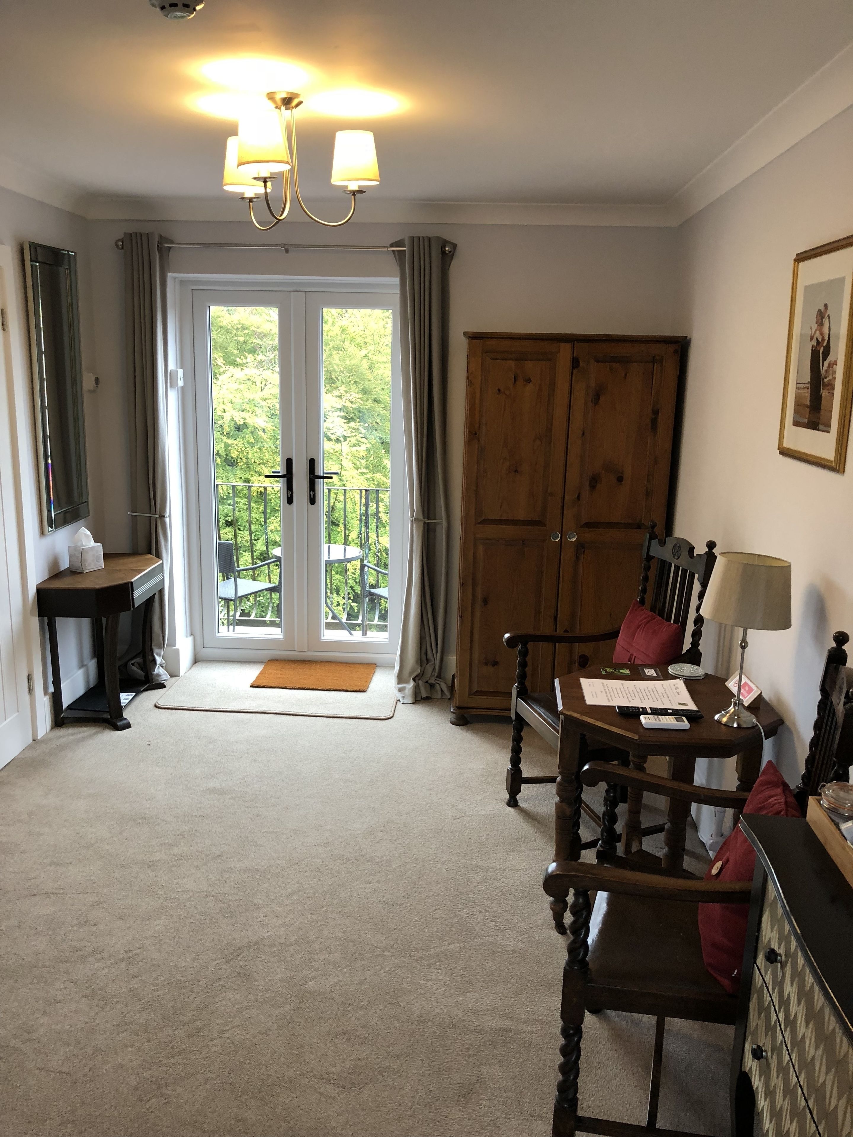 A room with a wooden wardrobe, a desk with a lamp, a chair, a small table, and a balcony door.
