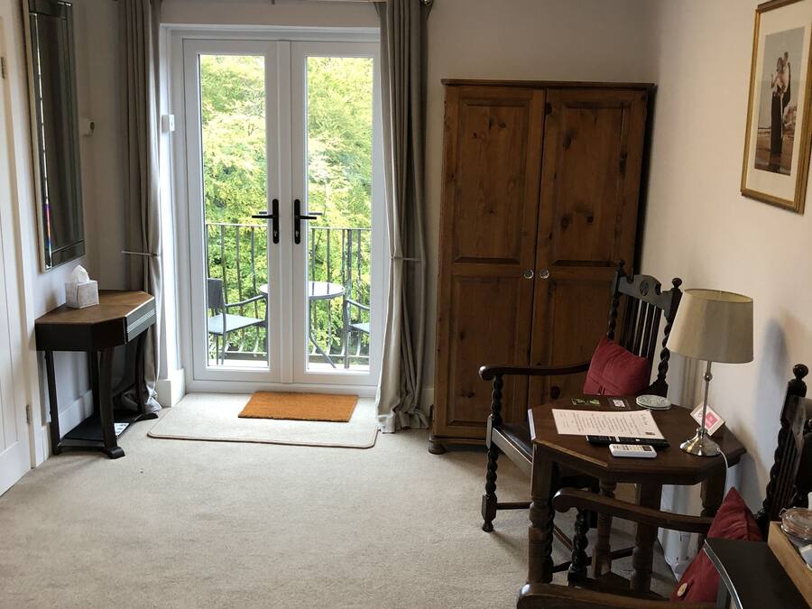 Una habitación con un armario de madera, un escritorio con lámpara, una silla, una mesita y una puerta al balcón.