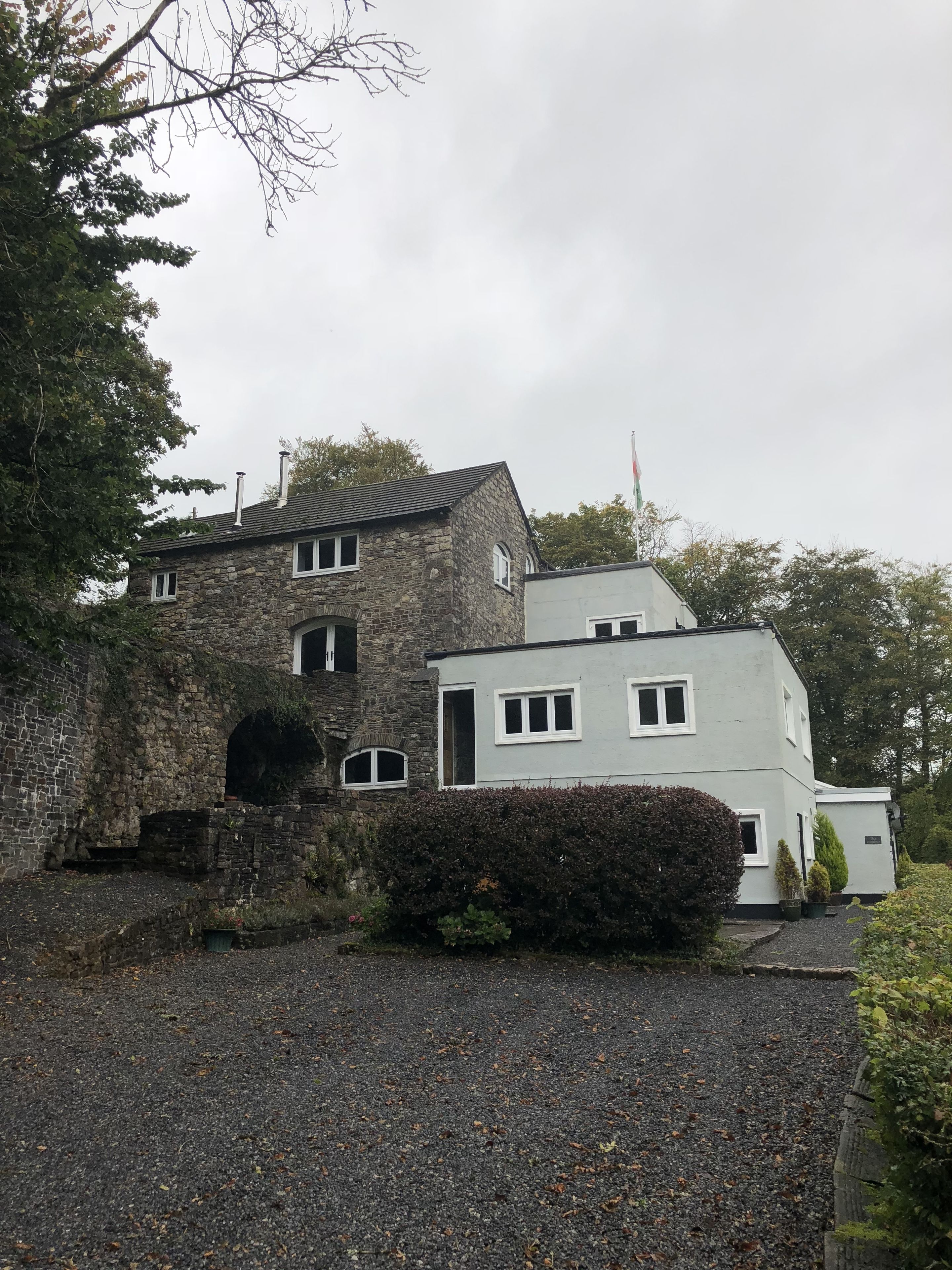 A stone building with a modern extension, a gravel driveway, and a hedge in front.