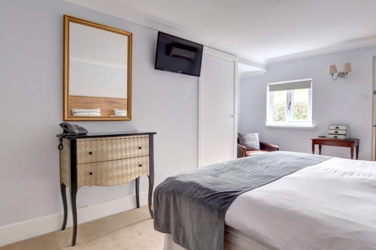 Une chambre à coucher comprenant un lit, une table de chevet avec une lampe, une commode, un téléviseur fixé au mur et une fenêtre laissant entrer la lumière naturelle.