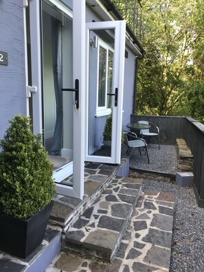 A stone-tiled patio with a small table and chairs, a potted plant, and a door leading to a covered outdoor area. - The Mill at Glynhir (Ammanford)
