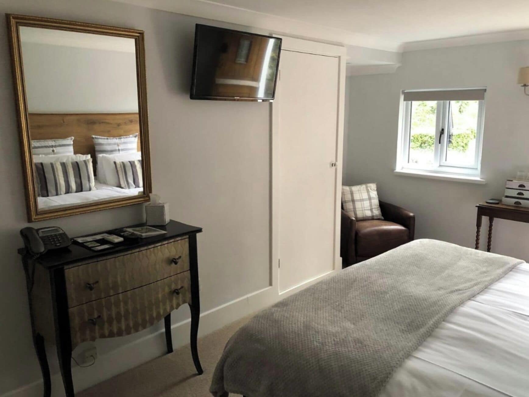 Un dormitorio con cama, escritorio con teléfono, televisor montado en la pared, armario y ventana con vista a la vegetación.