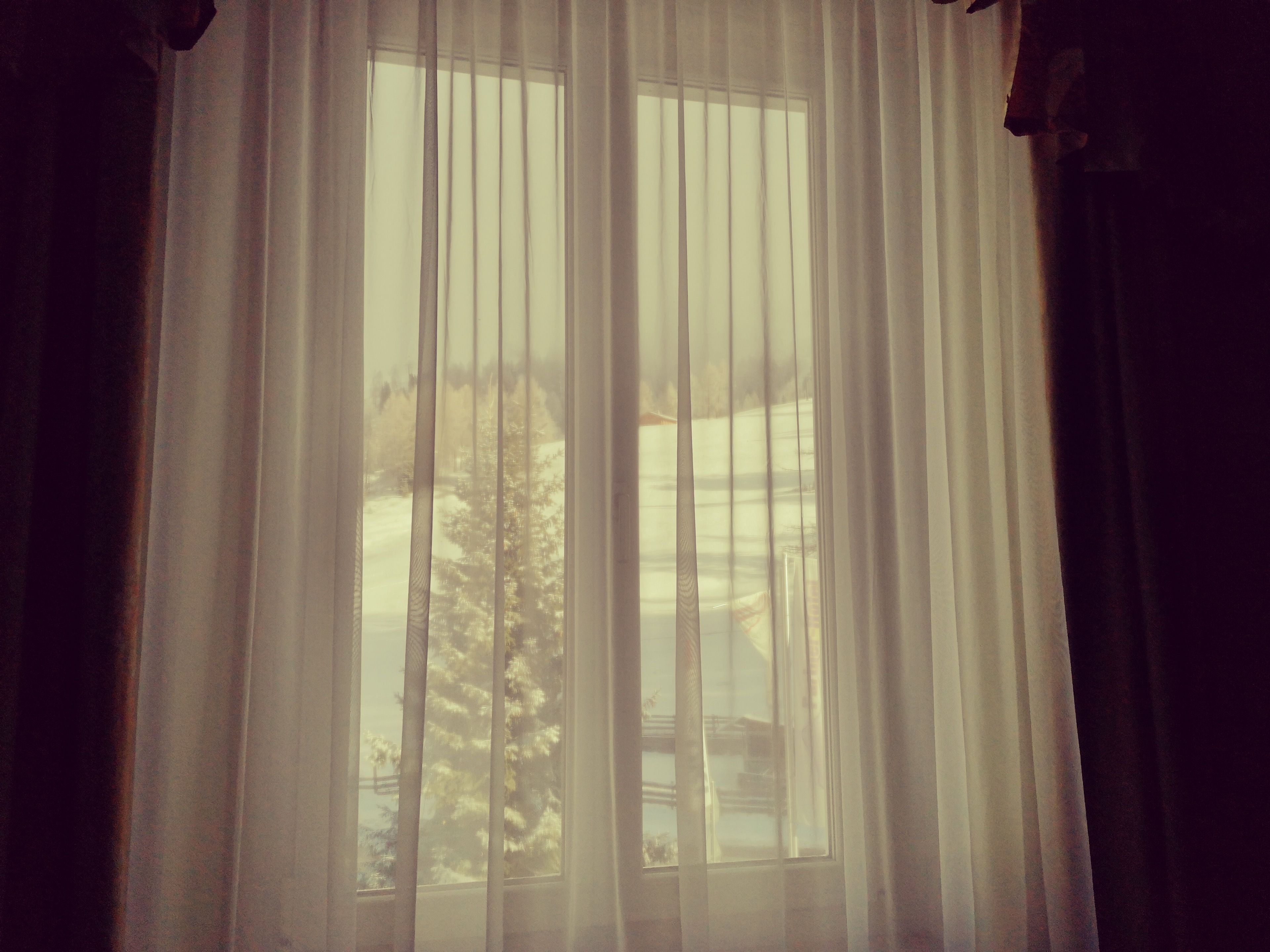 A window with sheer curtains, allowing a view of a snowy landscape and trees outside.