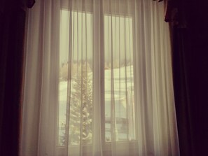 Ein Fenster mit leichtem Vorhang, durch den ein Blick auf eine verschneite Landschaft und Bäume ist.