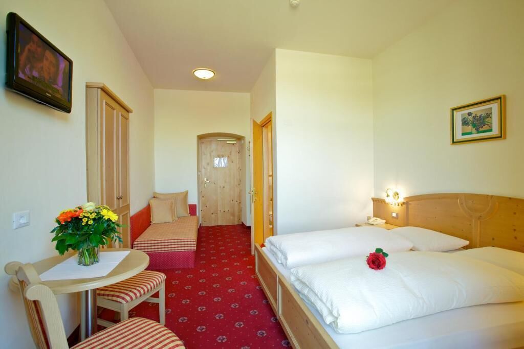 Een hotelkamer met een bed, een bureau met een stoel, een klein tafeltje met een bloevas en een deur naar een andere kamer.