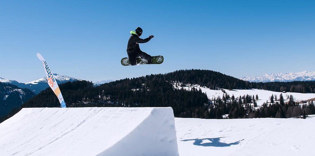 A snowboarder performing a jump over a snow ramp with a clear view of snow-covered mountains in the background.