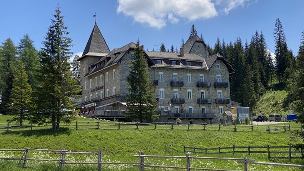 Un gran edificio de piedra con varios balcones y un techo a dos aguas pronunciado, rodeado de un exuberante paisaje verde y árboles altos.