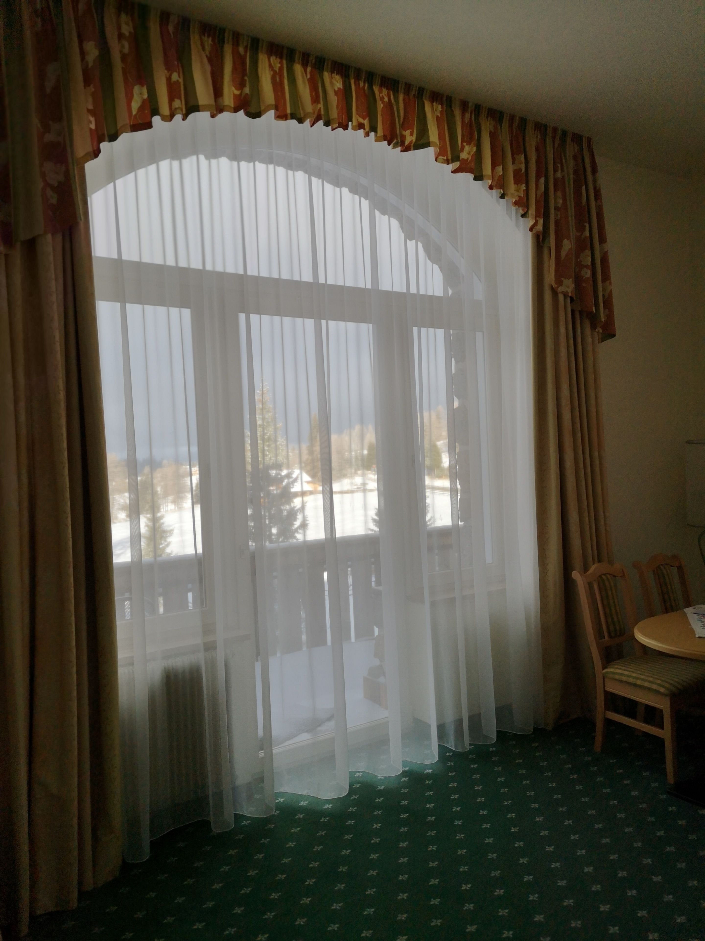 A room with a large window covered by sheer curtains, a wooden chair, and a small table.