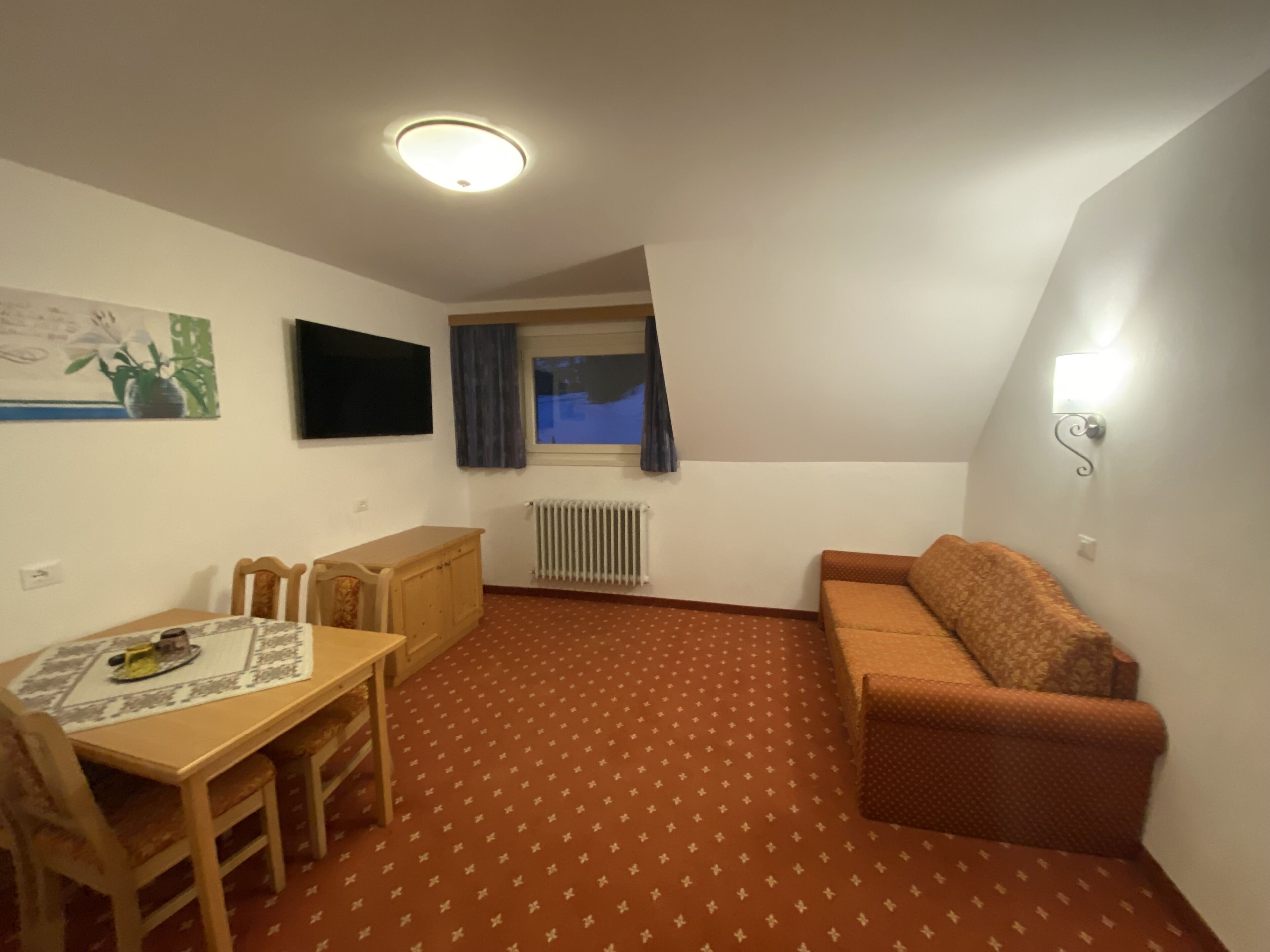 A small, cozy room with a dining table, chairs, a sofa, a TV mounted on the wall, and a window with curtains.