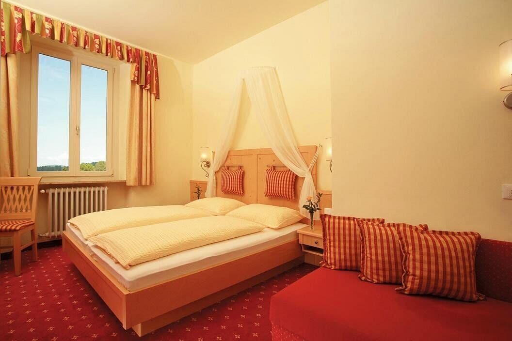 A bedroom with a large bed, a window with curtains, a bedside table with a lamp, and a red sofa.