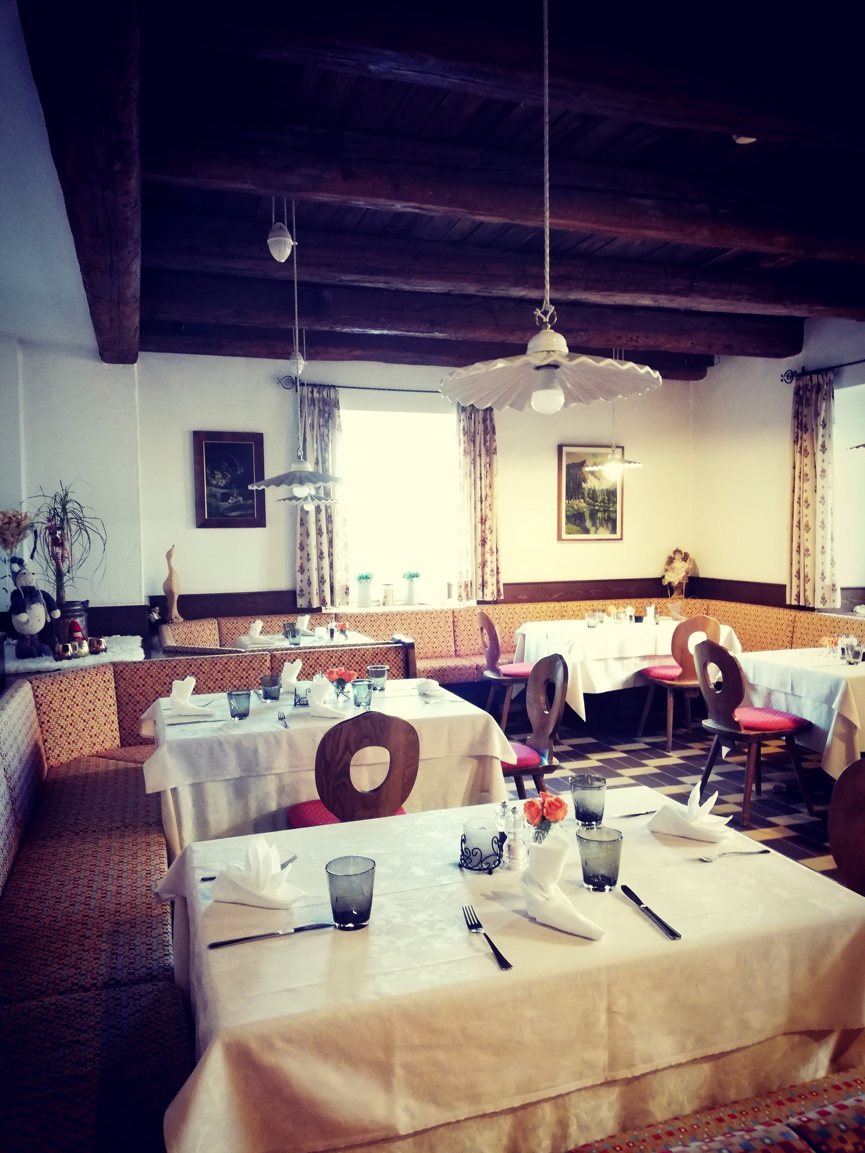 A restaurant with multiple tables and booths, wooden beams on the ceiling, and large windows with curtains.