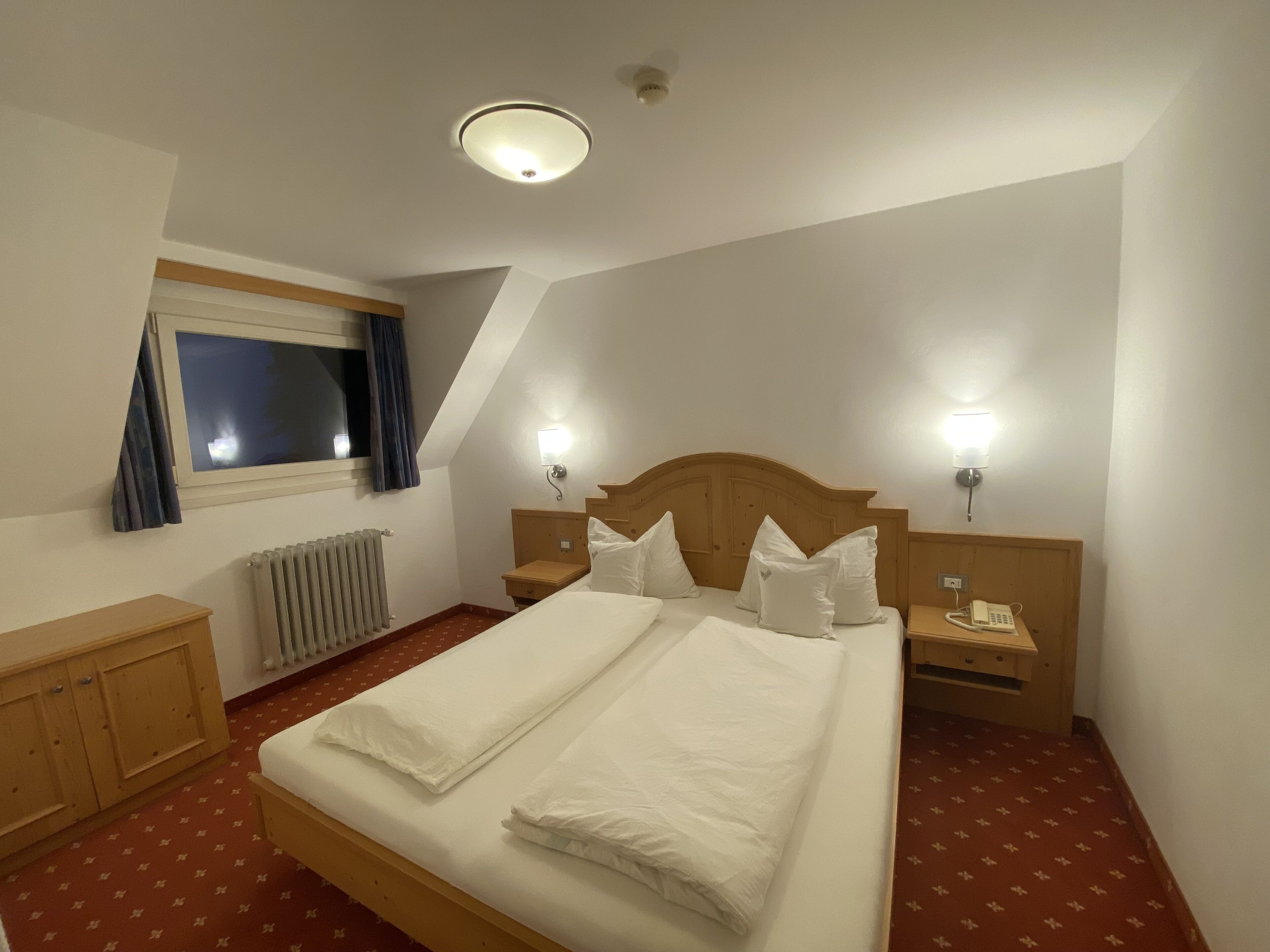 Ein Hotelzimmer mit einem großen Bett, zwei Nachttischen, einem Holz-Kopfteil, einem Fenster mit Vorhängen, einem Heizkörper und einem Telefon auf einem der Tische.