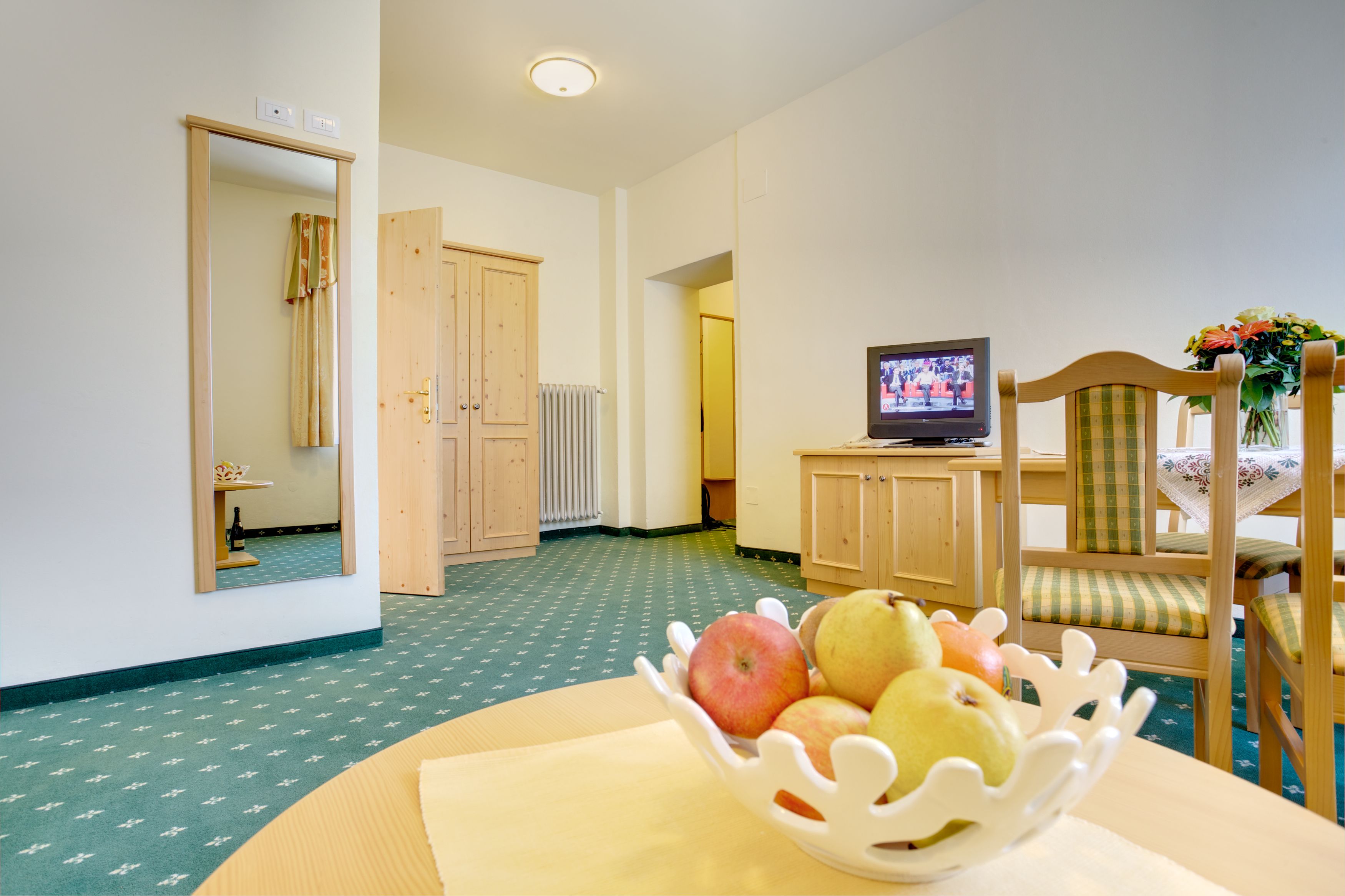A room with a wooden cabinet, a television, a chair with a checkered cushion, and a table with a white fruit bowl.