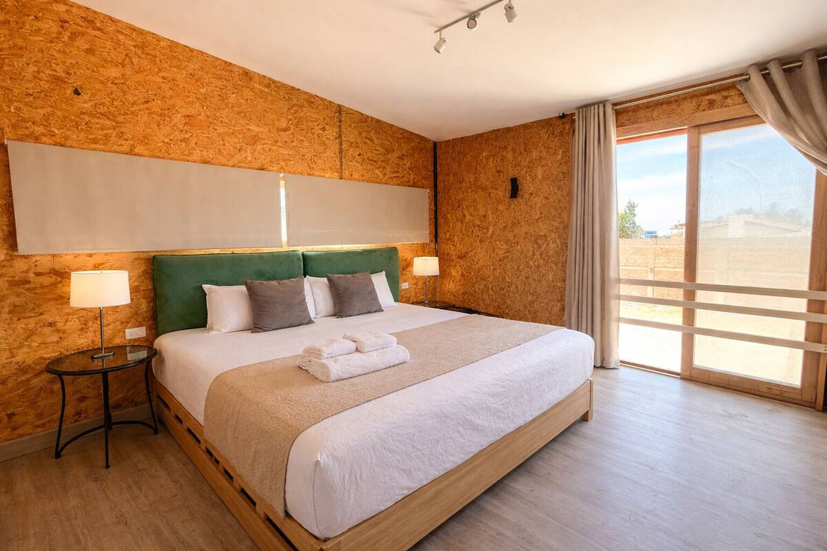 A hotel room with a large bed, wooden headboard, two bedside lamps, and a view through a large window.
