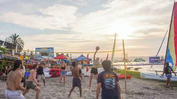 Partido de voleibol de playa con una red, palmeras y gente en el fondo.
