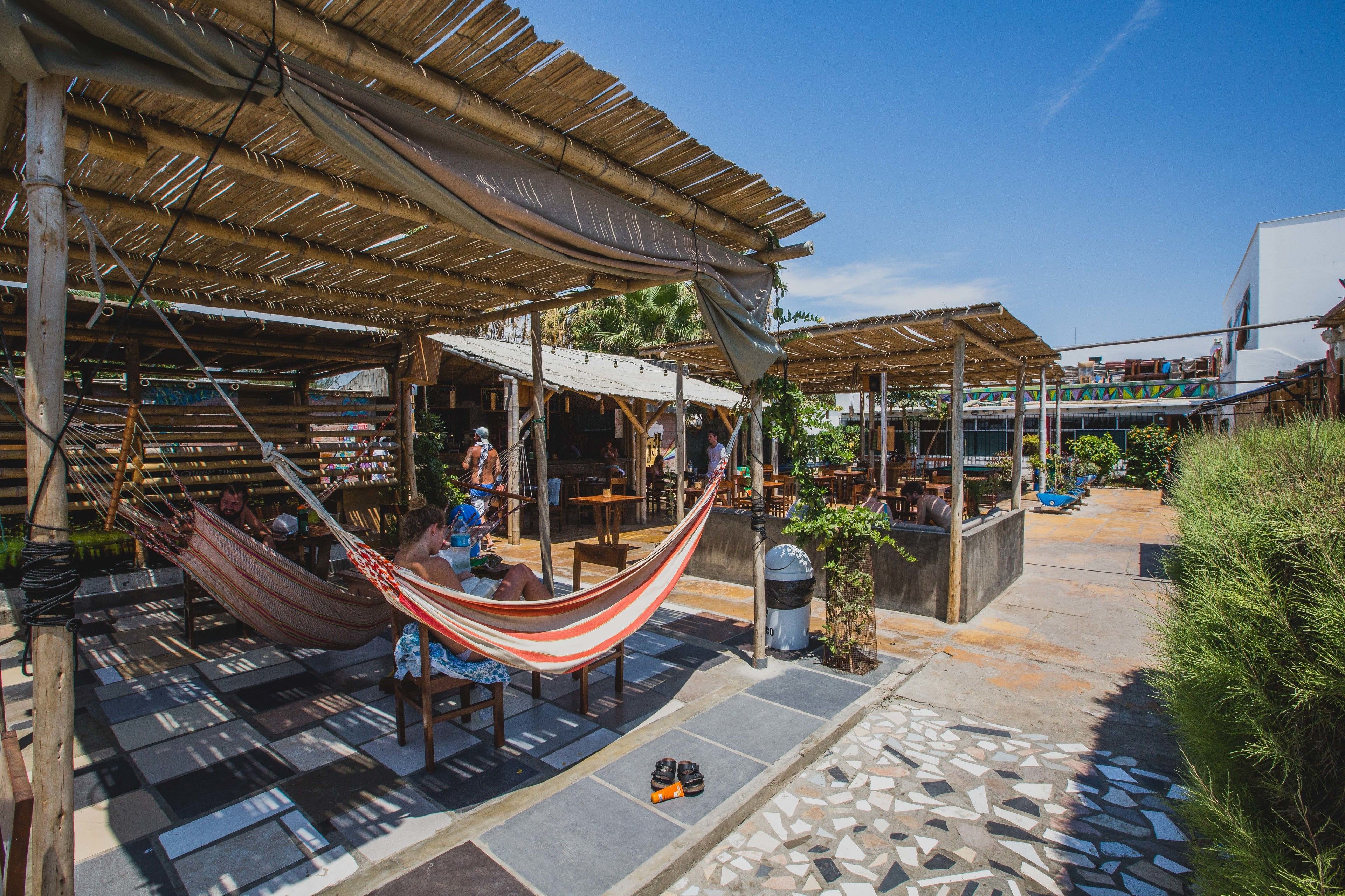 An outdoor seating area with hammocks, wooden tables, and chairs under a thatched roof.