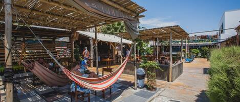 An outdoor seating area with hammocks, wooden tables, and chairs under a thatched roof.