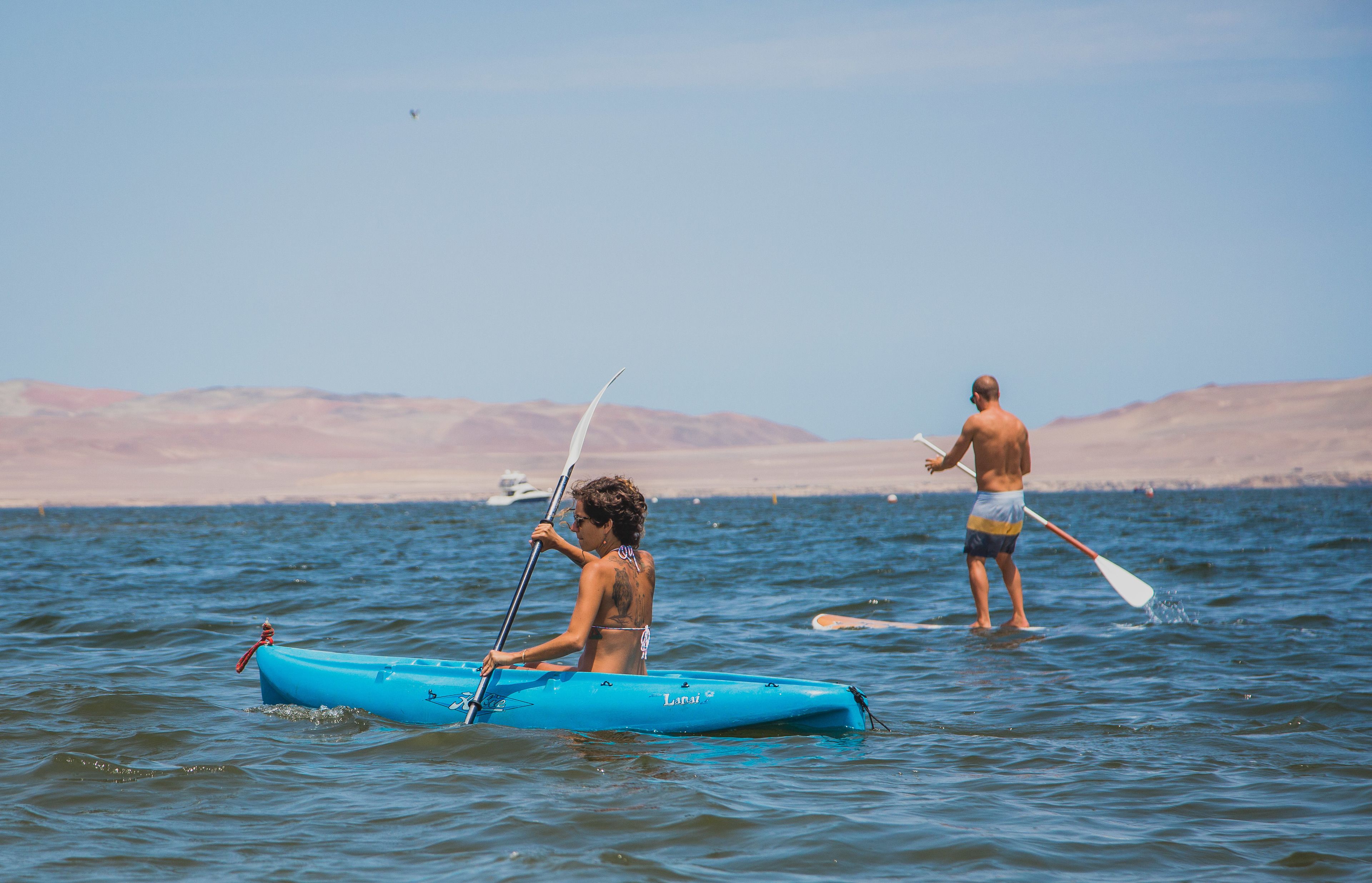 Two people in a blue kayak on calm water with a clear sky and distant mountains.