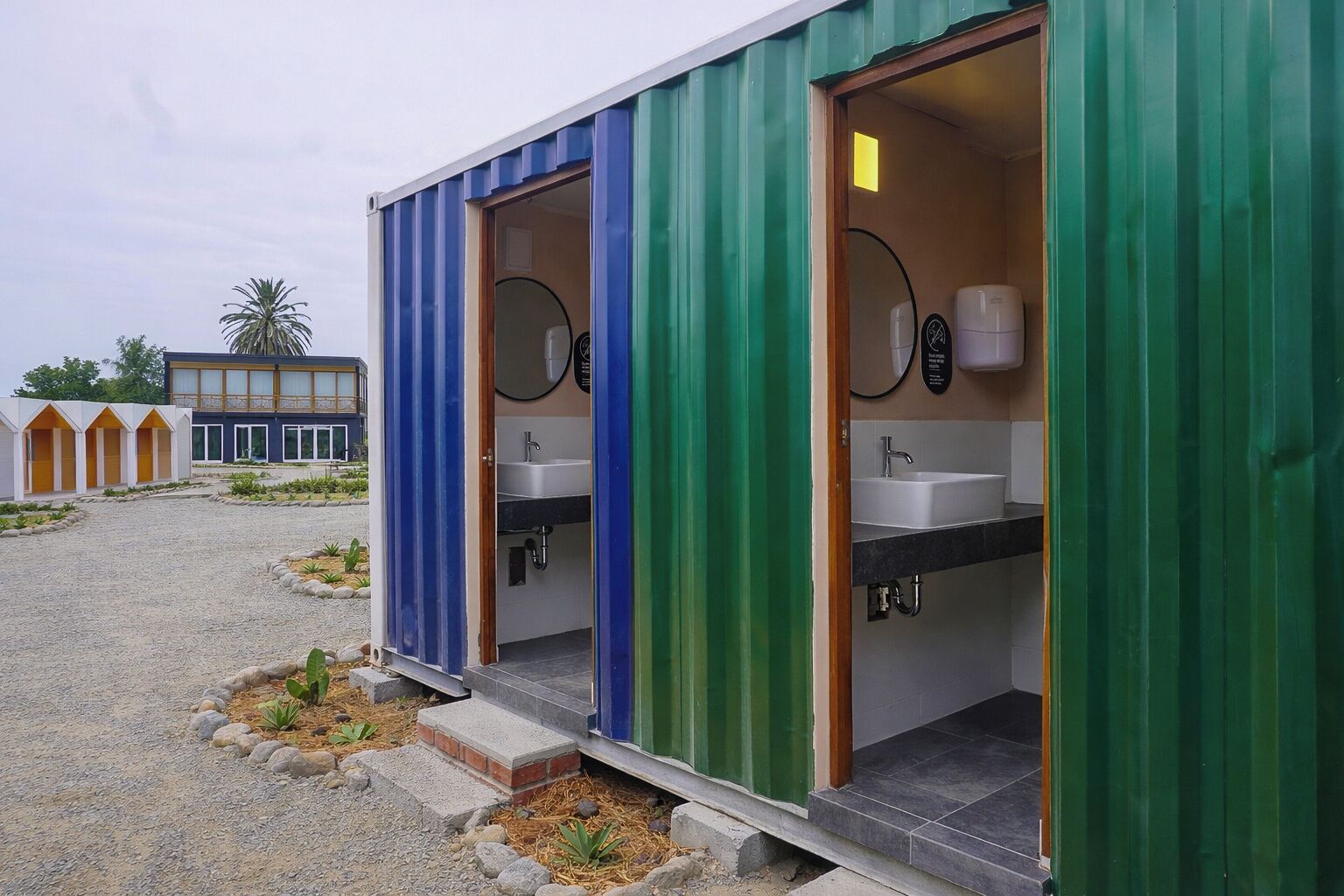 Portable restrooms with sinks and mirrors, surrounded by a gravel area and small plants.