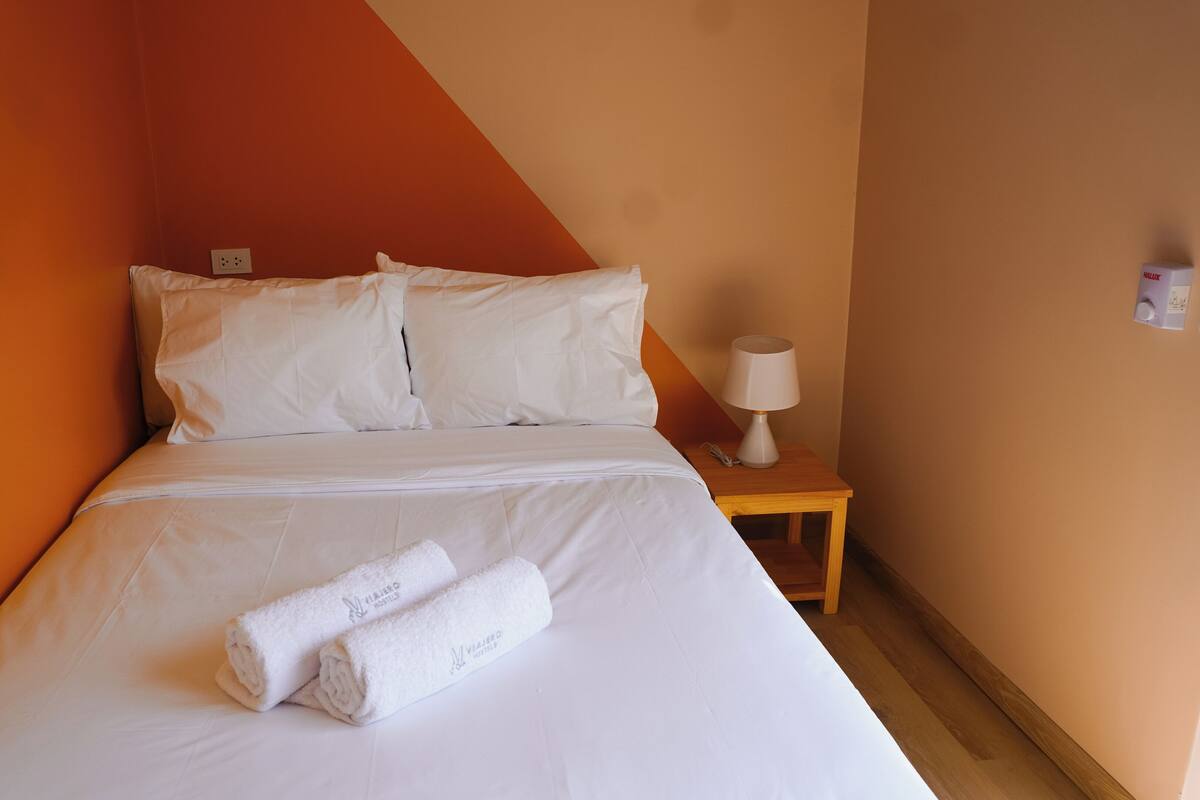 A single bed with white linens, a wooden nightstand with a lamp, and a rolled towel on the bed.