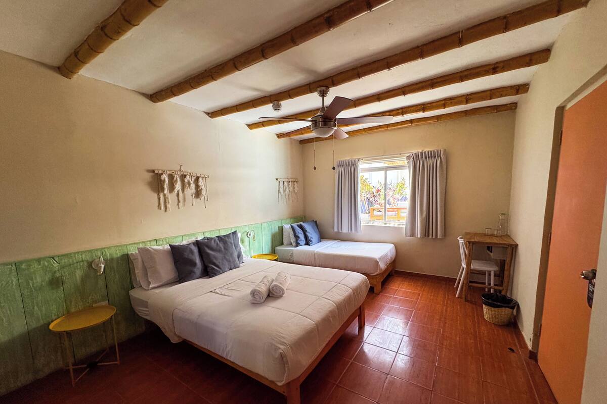 A room with two beds, a ceiling fan, a window with curtains, and a small table with a chair.