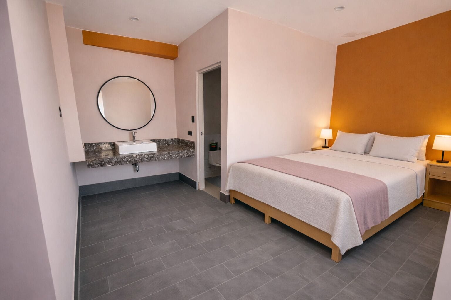 A hotel room with a bed, bedside table with a lamp, a round mirror above the sink, and a door leading to a bathroom.