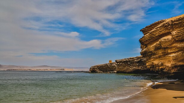 A coastal landscape with a sandy beach, clear blue water, and a rocky cliff. - Viajero Paracas Hostel (Paracas)