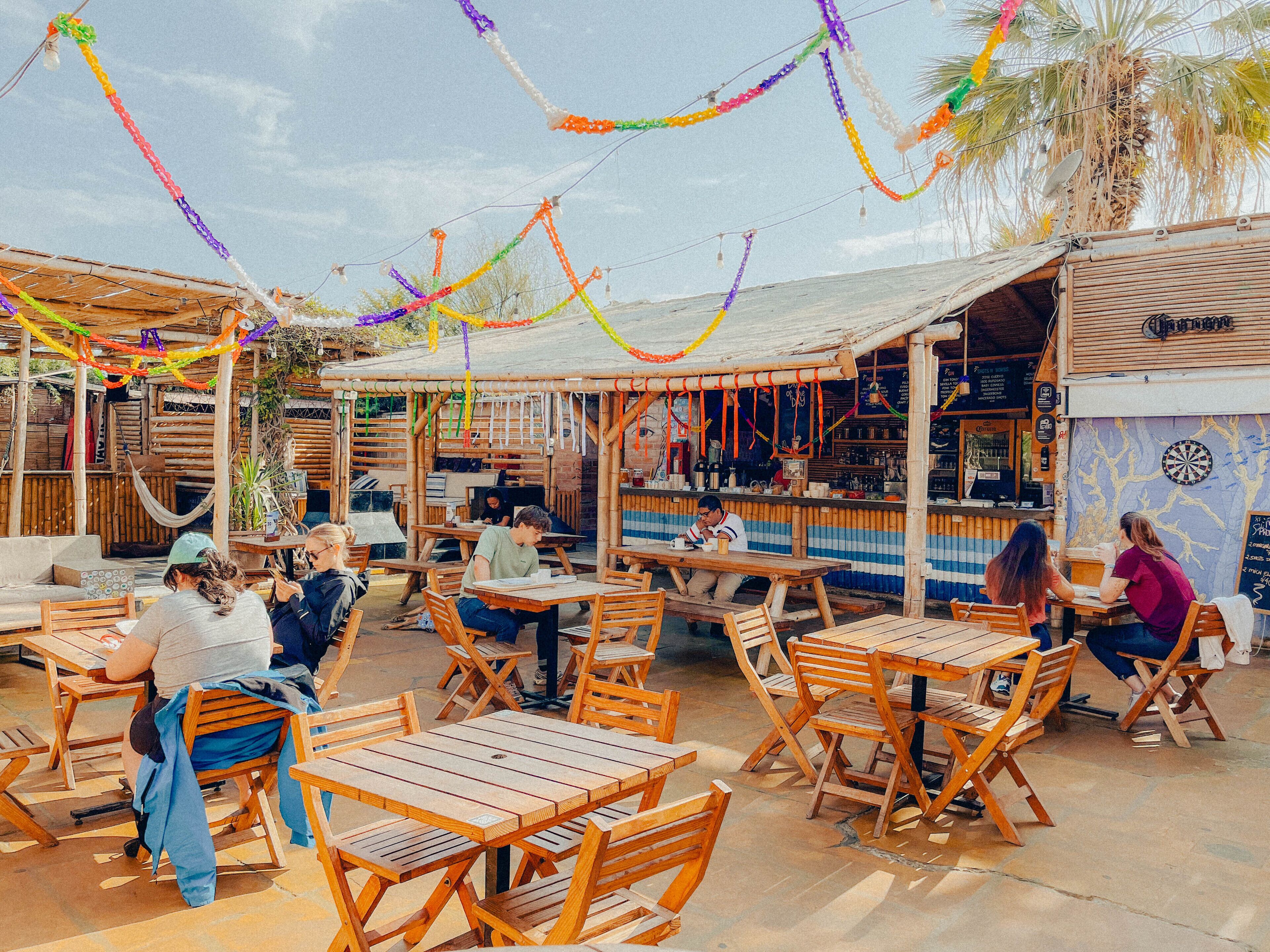 An outdoor seating area with wooden tables and chairs, a bar with a dartboard, and a sign for 'The Bar'.
