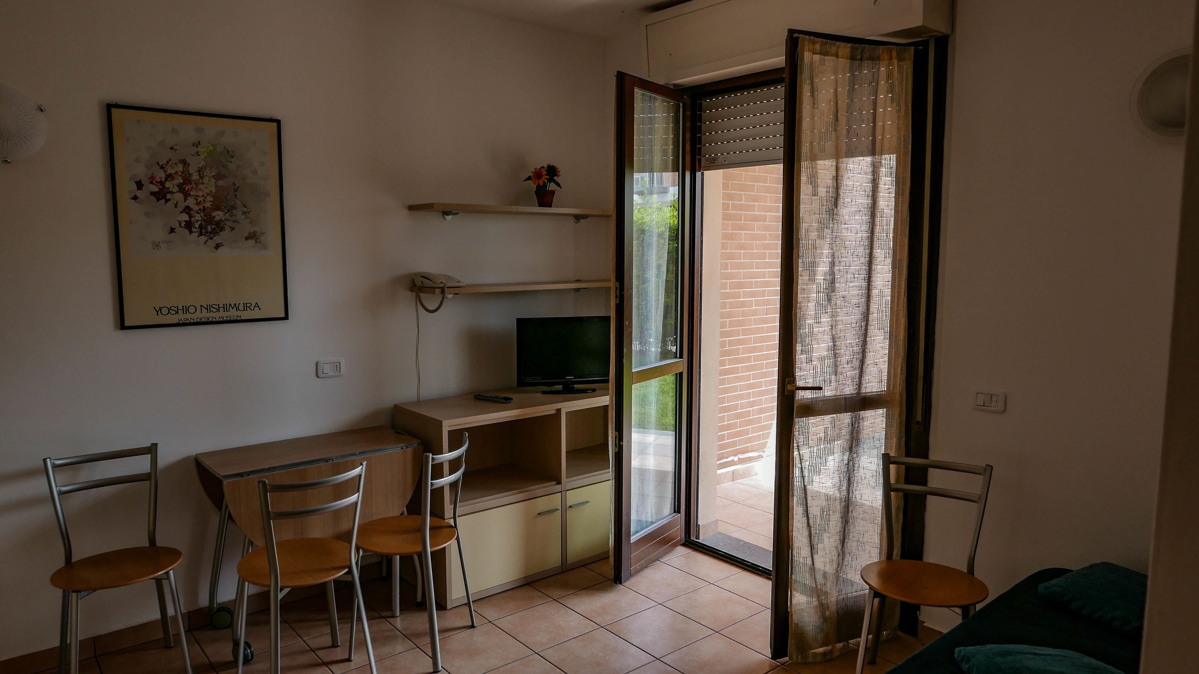 A small, open-plan living space with a desk, chairs, a television, and a sliding glass door leading to a balcony.