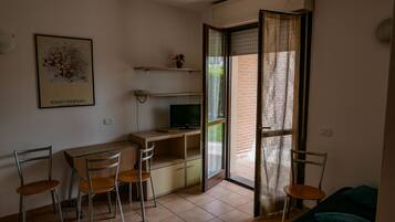 A small, open-plan living space with a desk, chairs, a television, and a sliding glass door leading to a balcony.