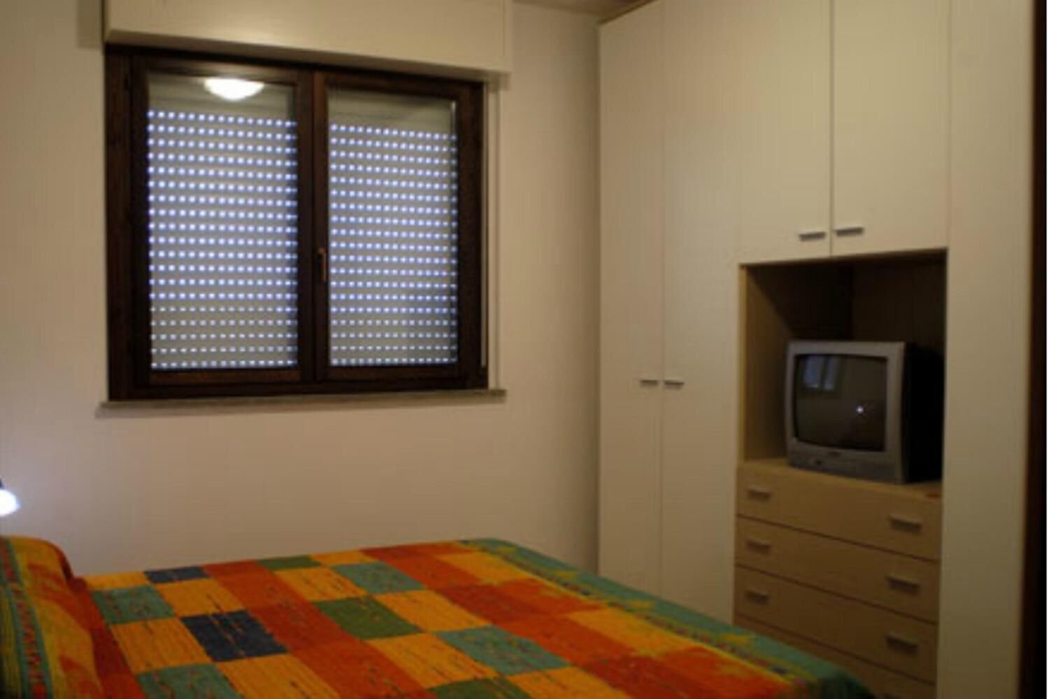 Ein Schlafzimmer mit einem Bett, einem Fenster mit Jalousien, einem Fernseher auf einem Schrank und einem Kleiderschrank.