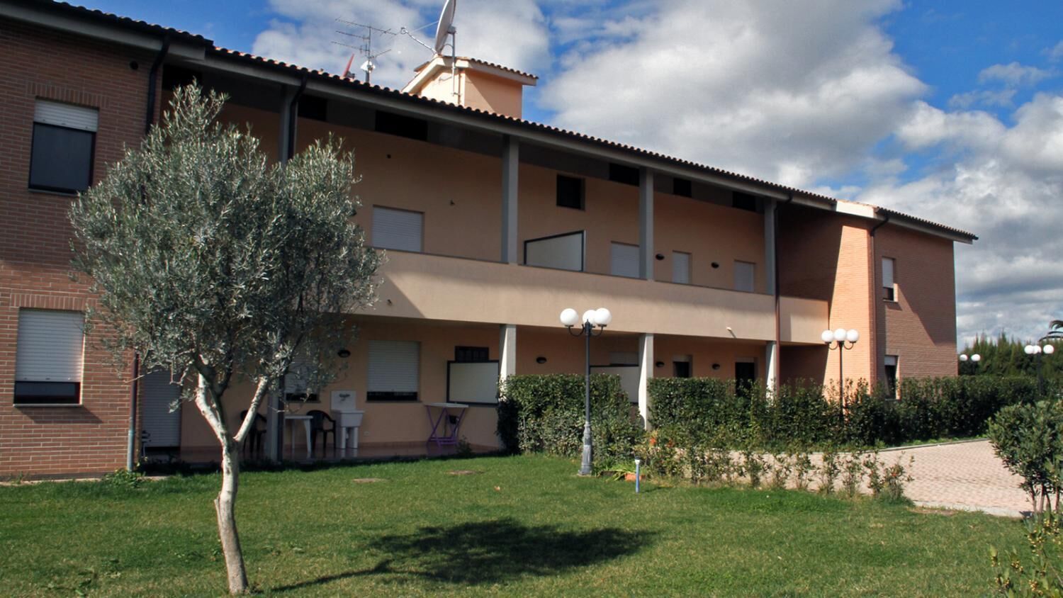 Un edificio di due piani con tetto piatto, un’antenna parabolica e un piccolo albero nel giardino antistante.