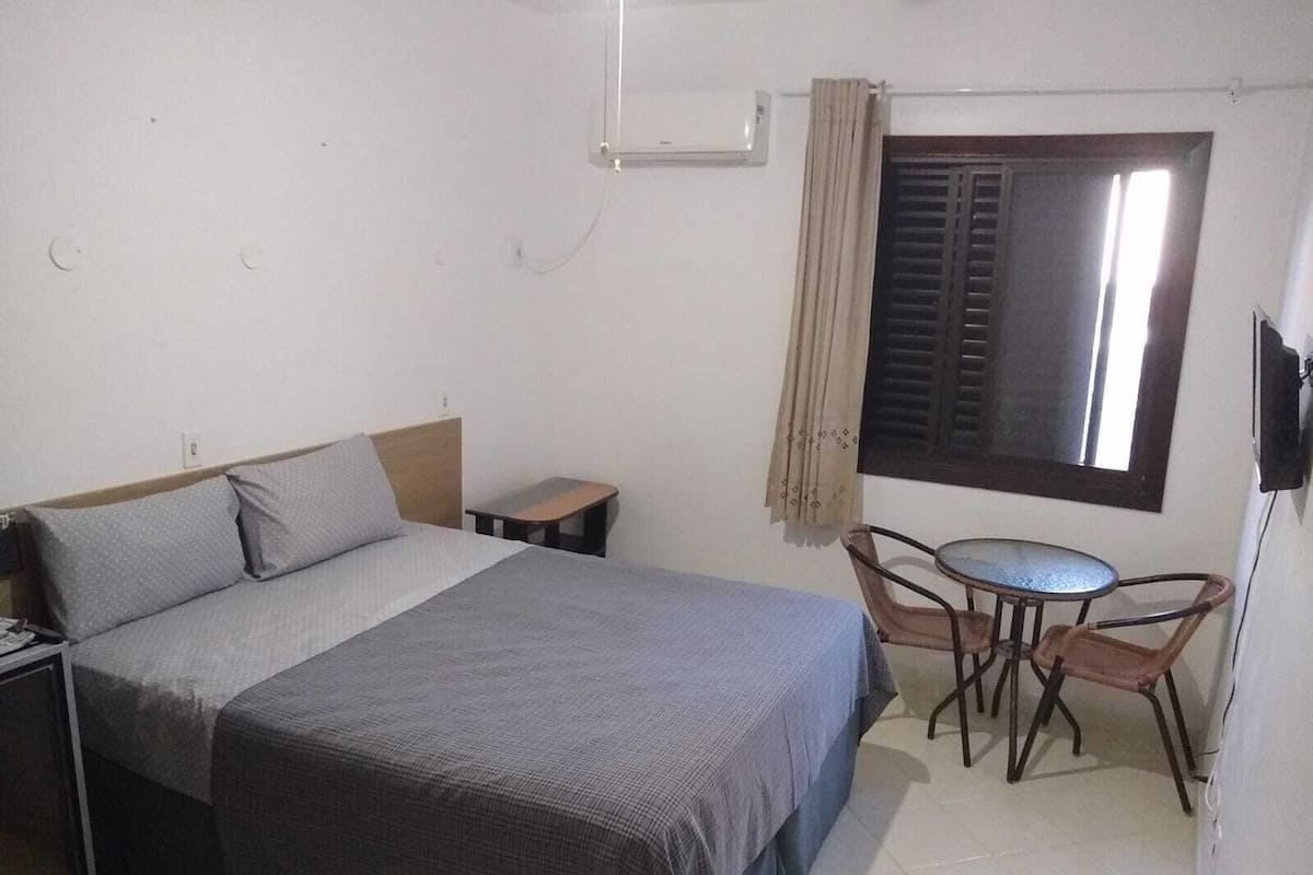 A bedroom with a bed, a small table, a chair, a window with shutters, and a wall-mounted TV.