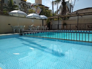 A swimming pool with tiled flooring, surrounded by white plastic chairs and umbrellas. - Parque Suítes Guarujá (Guarujá)