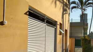 Un edificio con una puerta persiana cerrada, un camino empedrado y un pequeño jardín con plantas y una fuente.