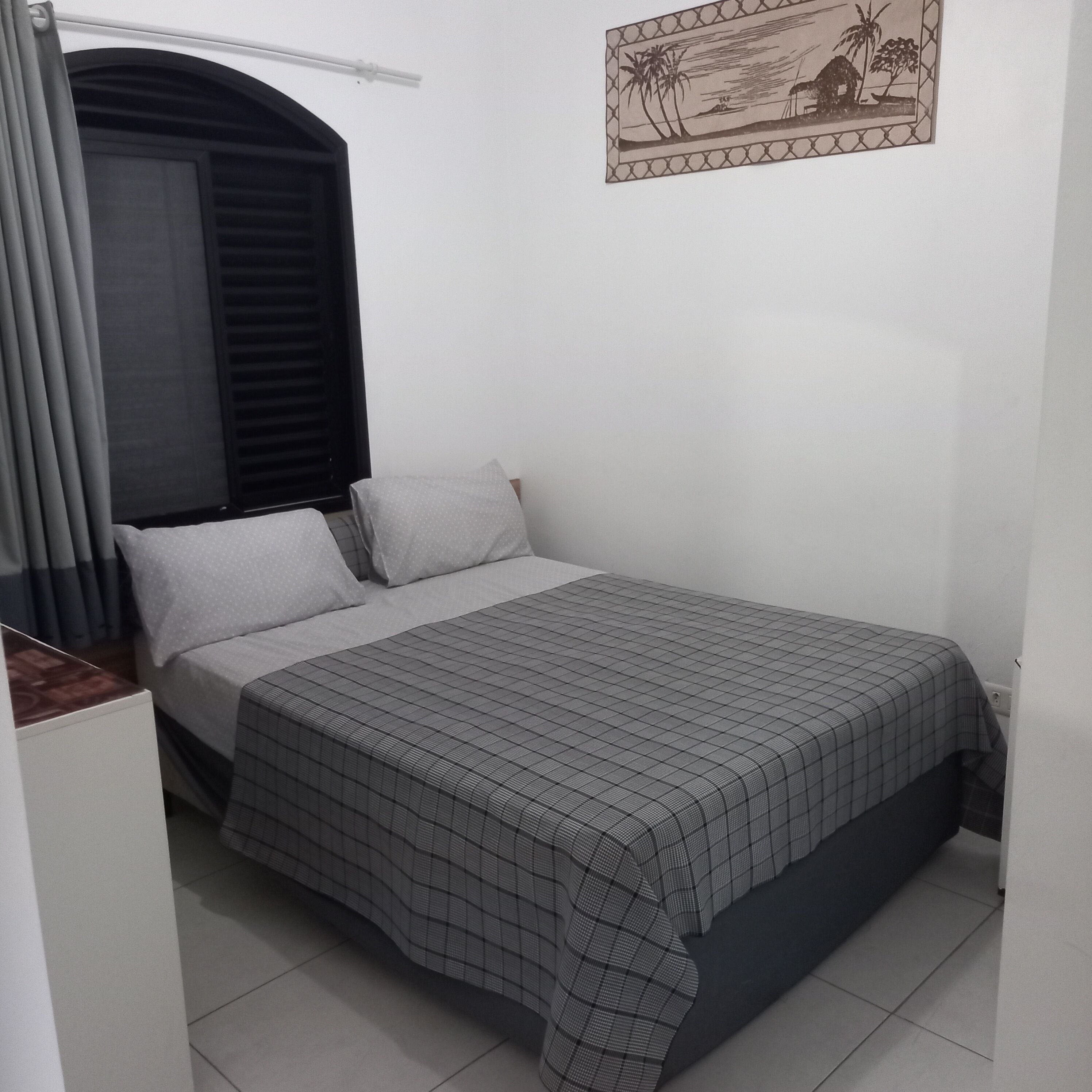 A bedroom with a bed, curtains, a window with blinds, a wall-mounted picture, and a tiled floor.