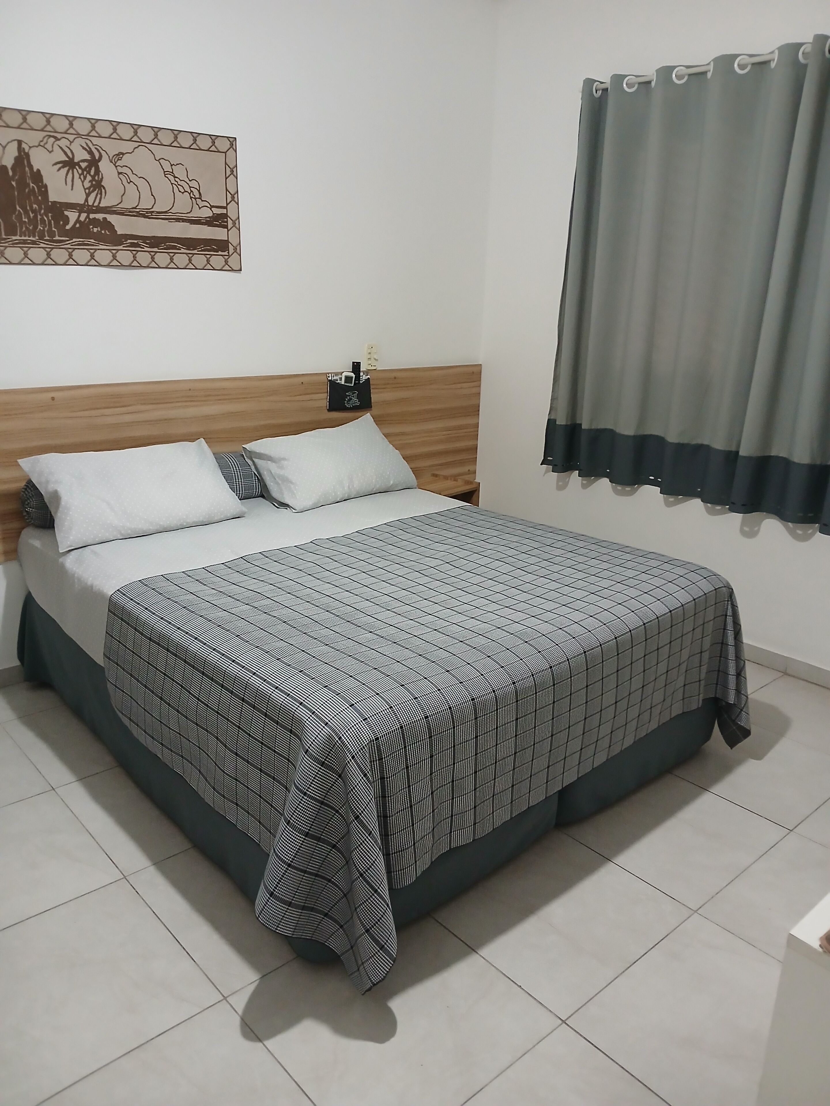 Un lit bien fait, avec une couvre-lit à carreaux, une tête de lit en bois et une table de chevet sur laquelle se trouvent une lampe et un petit objet décoratif.